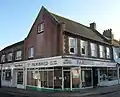 Three-quarter view of a two-storey red-brick and cobbled flint building on a corner site. The ground floor is occupied by four shop units, of which a café called "Famished" is most prominent. At first-floor level, the right-hand face has five evenly-spaced windows with red-brick surrounds. The gable end of the roof, which forms the larger part of the left-hand face, is blank apart from two small windows. Beyond this, there is a three-window range in a wall of dark brickwork at first-floor level.