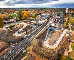 A train station in a small town, as seen from a drone, showing footpaths leading to platforms on both sides of the track. A station house is to one side.