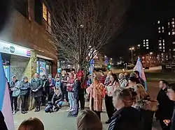 A group of people gathered in a circle on a sidewalk at night. Many are holding transgender pride flags.