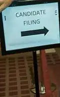 A sign with a black frame holds a piece of paper reading "CANDIDATE FILING" with an arrow pointing to the right