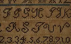 Close-up of an alphabet embroidery sampler made with blue, green, black, and yellow thread. The rows of letters are each separated by a line of herringbone stitches.