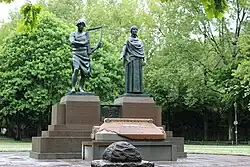 two bronze statues on pedestals, against a background of trees