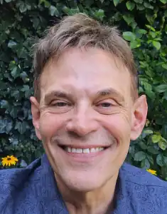 Eric Heinze wears a blue shirt and smiles widely, He is standing outside in front of a green hedge dotted with yellow flowers.