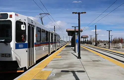 A white train is parked at a recessed side platformed station with canopies and signage visible.