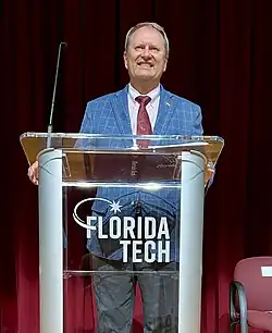 Provost John Z. Kiss in blue blazer and crimson tie standing at acrylic Florida Tech podium.