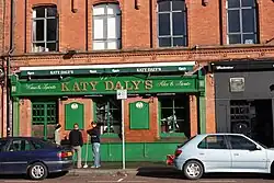 Green-fronted Irish pub, with "Katy Daly's" written on the hoarding above. A black-fronted door is to the right, and cars are parked on the roadway in front.