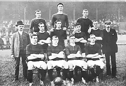 A black-and-white photograph of a football team lining up before a match. Four players, wearing dark shirts, light shorts and dark socks, are seated. Four more players are standing immediately behind them, and three more are standing on a higher level on the back row. Two men in suits are standing either side of the players.