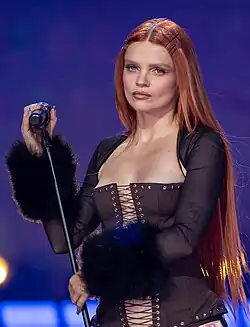 Redhead female in a brown corset top holding a microphone on a stand as she looks into the distance.