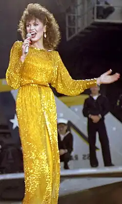 A young woman with long brown hair, wearing a long yellow dress, singing into a microphone