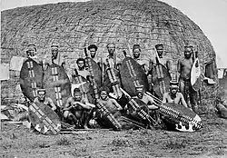 African men in loin cloths with spears and cowhide shields standing in front of a grass hut