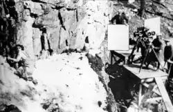 Nell Shipman hanging off of a cliff while Joseph B. Walker films the scene and Clif Maupin holds a reflector.