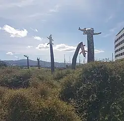 clear blue sky, foreground are plants, and long thin bending mokomoko sculptures with flapping arms