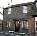 Close-up view of a two-storey, four-window, semi-detached cottage in dark, irregular stone with red brickwork around the walls, windows and door. There is a flat shelf-like white structure above the black door. A white-painted chimney sits on the left side of the roof.