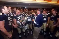 A middle aged man in a blue polo shirt and khaki pants speaks to young men in football uniforms in the locker room. One of the men is holding a trophy.