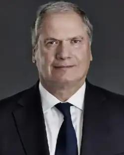 stocky caucasian male, 60s, with thinning grey hair wearing a dark suit, white shirt and black tie