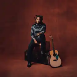 Shaboozey looking ahead and sitting on two stacked trunks with an acoustic guitar by his side.