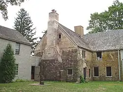 The rear of a 2+ story stone house has a wooden addition extending off to the left. An old-fashioned water pump with bucket is near the door.