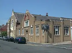 Three-quarter view of a brownish building on a corner site, with cars parked in front of its main façade. This has a single storey with two taller gabled sections, the larger of which (furthest away in the picture) has a short tower-style structure next to it. A black and white sign hanging from the corner is headed "Stoneham Road Baptist Church".