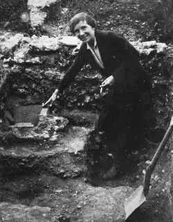 Photograph of a woman in a long coat, standing in an archaeological trench. She points at something on the ground; a shovel rests next to her.