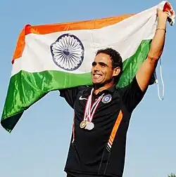 Picture of a young Indian male up to the waist. He is holding India's national flag over his head and with medals around his neck