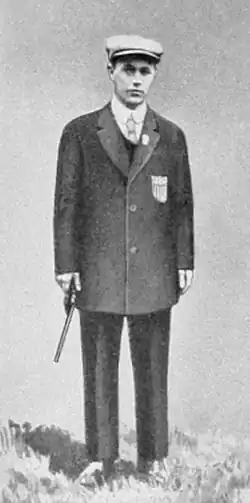 Alfred Lane standing with a gun by his side. He's wearing a sports coat, slacks, and a hat.
