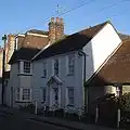 Three-quarter view of a white-painted cottage with a brown tiled roof, three first-floor windows and a pedimented doorcase flanked by two windows on the ground floor. It is attached to a pale yellow building on the left; this is slightly taller and has square corner windows. To the right, a single-storey building with a long sloping roof shares a wall.