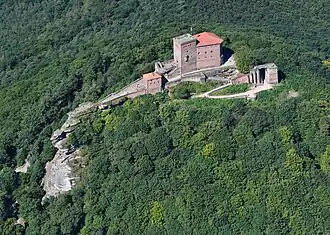 Photo of Trifels Castle as seen from the air. The Castle's square brick towers sit on top of the rocky peak of a hill in a forested valley.