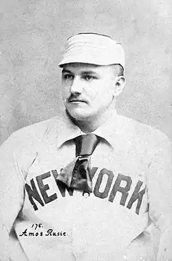 A seated man in a white baseball cap and white baseball uniform shown from the chest up. He is looking to the left of the image. He has a small mustache, and his jersey reads "NEW YORK" in dark block type, partially obscured by a short tie worn around his neck under the collar of his jersey.