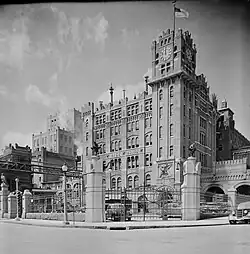 A towering, fenced in building with a clock tower and flagpole on one corner, in black-and-white.