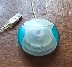 Overhead view of a round, translucent plastic peripheral on a wooden desk. Circuitry inside is partially visible beneath the blue-and-white plastic