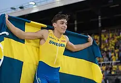 Photo of Armand Duplantis wearing a yellow and blue athletics outfit and holding up the yellow and blue Swedish flag behind his back