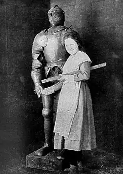 Black and white film still showing a woman standing next to a suit of armor