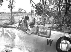 An old man wearing a hat and white shirt sitting in the cockpit of a silver aircraft. A younger man is leaning into the cockpit.