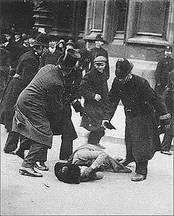 Black and white image of a woman in a light coloured dress and dark hat lying on her side in the street with her hands over her face, while several men stand in threatening postures above her.