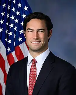 Official House portrait of Gill smiling in front of the U.S. flag, wearing a black suit, white shirt, and red tie with small white birds.
