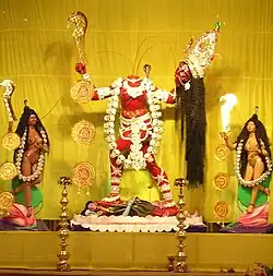 A decapitated, nude, red-complexioned goddess stands on a copulating couple inside a lotus. She holds her severed head and a scimitar. Three streams of blood from her neck feed her head and two nude (one fair, another brown coloured) women holding a scimitar and a skull-cup, who flank her. The goddess wears a skull-garland, a serpent (around her neck) and various ornaments.