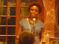 A Black woman with short, curly hair speaks into a microphone in a warmly lit room. She is wearing a black and white striped dress and layered necklaces. Behind her are wooden-framed windows and decorative cobwebs on the wall. A water pitcher sits on the table in front of her, and there are people in the audience.