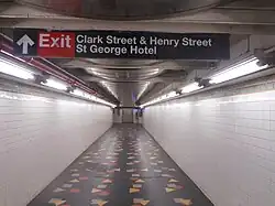 A passageway above the platforms, leading to the elevators. The passageway has black floors with multicolored shapes, which are part of an artwork. The walls are clad with white tiles. There is a sign on the ceiling, directing passengers to the station's exit in the St. George Hotel.
