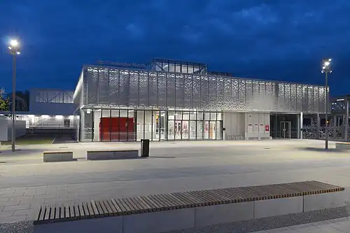 A large building with the facade made out of a metal mesh and large floor-to-ceiling windows on the ground floor. The photo is taken at night from behind a bench a short distance from the station.