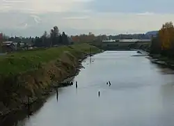 A stream perhaps 50 feet (15 m) wide flows between raised, grass-covered banks on either side. Several poles or sticks, perhaps remnants of a derelict structure, poke above the water in places. Low buildings and many trees are in the middle distance. A mountain range dominated by a snow-covered peak is visible in the far distance.