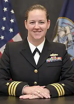 A white woman in a black US Navy uniform is sitting at a table with her hands folded upon it; she is smiling and looking into the camera.