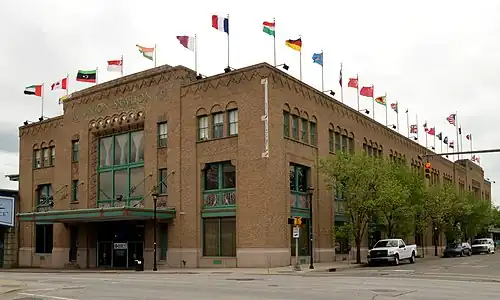 A large, rectangular, tan brick building with green trim and a roof lined with flags