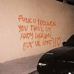 The album title spray painted in orange on a beige wall with a car in the foreground