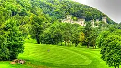 a golf course, in the background is the top of a castellated mansion can be seen within the woodland