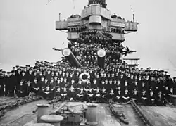 A large number of men posing for a photo on the foredeck of a warship. Two of the ship's gun barrels are visible in the middle of the group.