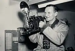 An image of Henry Busse in 1947. He's smiling and holding a flash bulb camera.