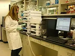 A woman in a lab coat takes glass vials out of a machine. A computer displays a spectroscopy graph.