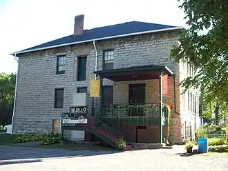A different side of the same stone building, with stairs and a metal porch to the right from which flowering plants hang. There are banners on the porch identifying it as the Jell-O Museum in Le Roy, New York