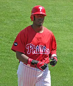 A dark-skinned young man wearing a red baseball jersey and batting helmet and white pinstriped baseball pants