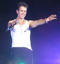 Joey McIntyre wearing a white tank top and dark pants, holding a microphone onstage, with arms outstretched toward audience